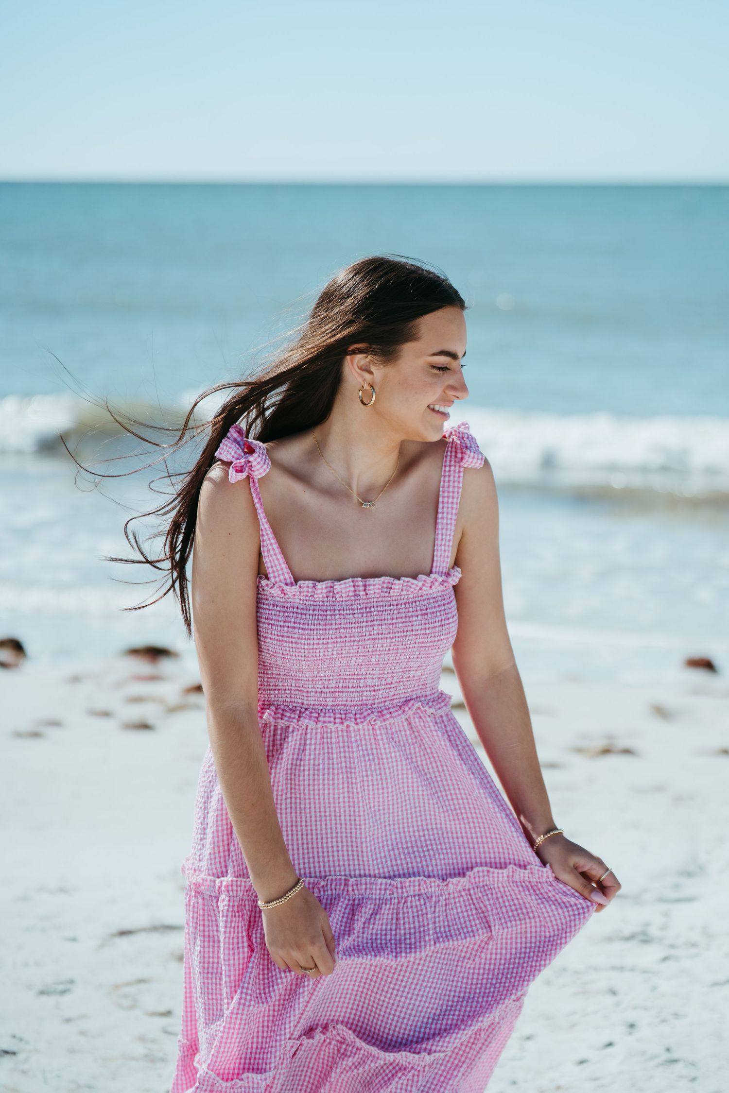 Woman in pink gingham midi dress smiling on a sunny beach, hair blowing in the wind.