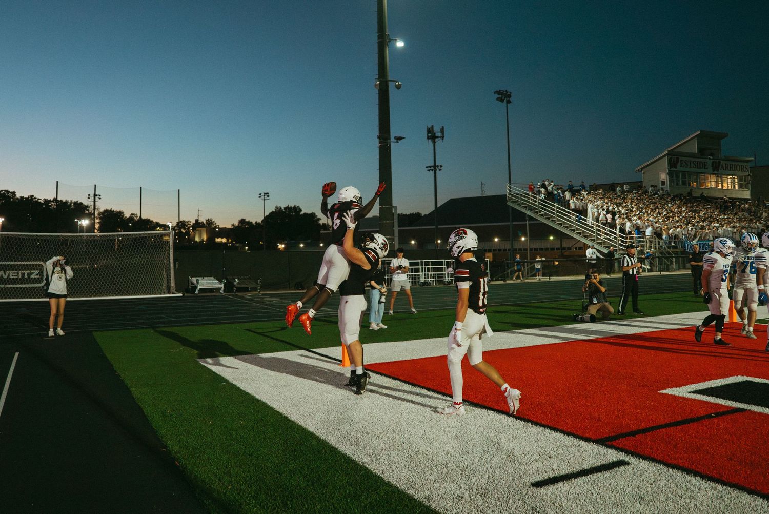 Football players celebrate a touchdown in the end zone under stadium lights at dusk on a red turf field.
