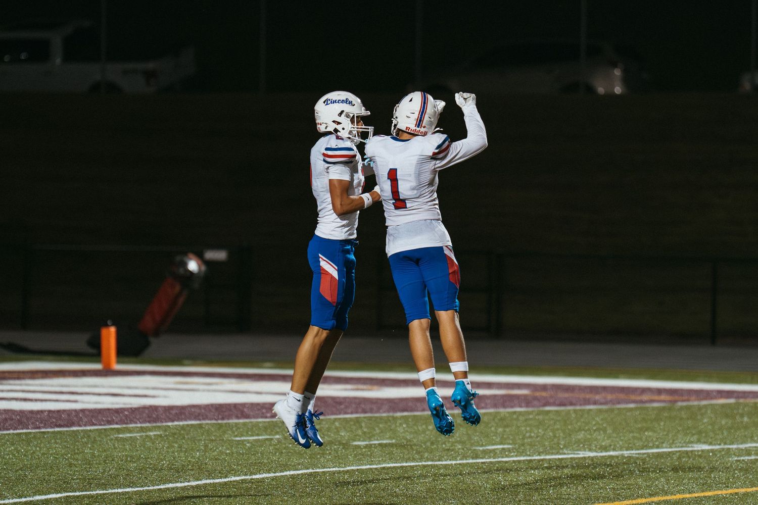 Two football players in white uniforms celebrate a touchdown on the field at night.