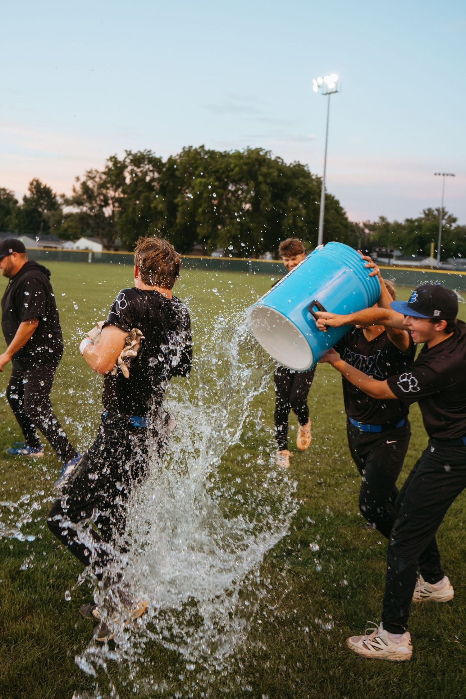 Baseball players celebrate victory as teammate gets drenched with blue bucket of water on field at dusk.