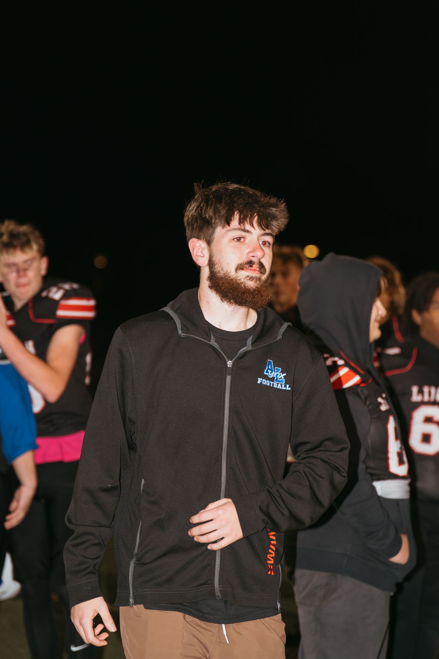 Young man with beard wearing black football zip-up jacket stands outdoors at night near football players.