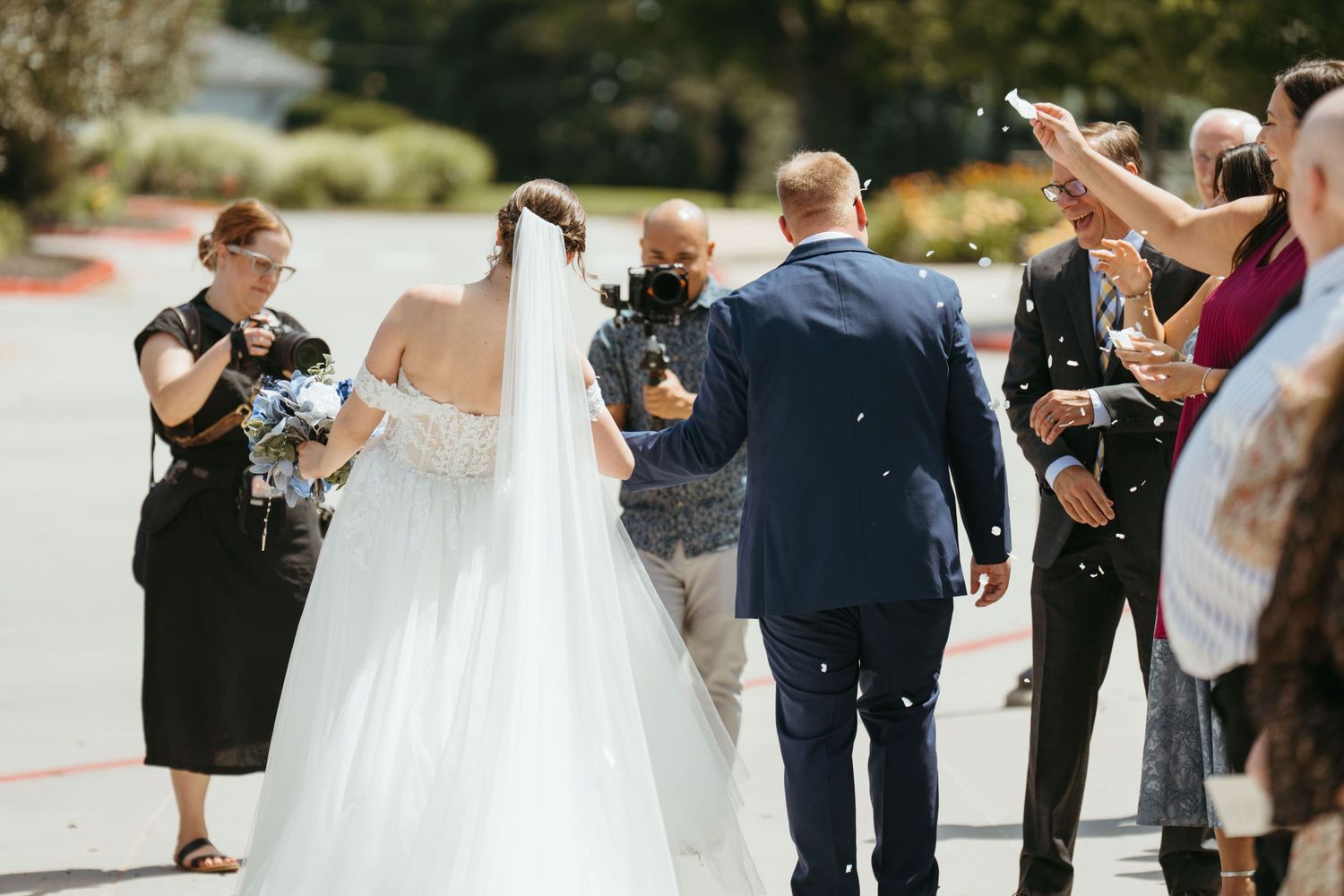 Bride and groom walk through confetti shower with wedding guests celebrating outdoors on sunny day.