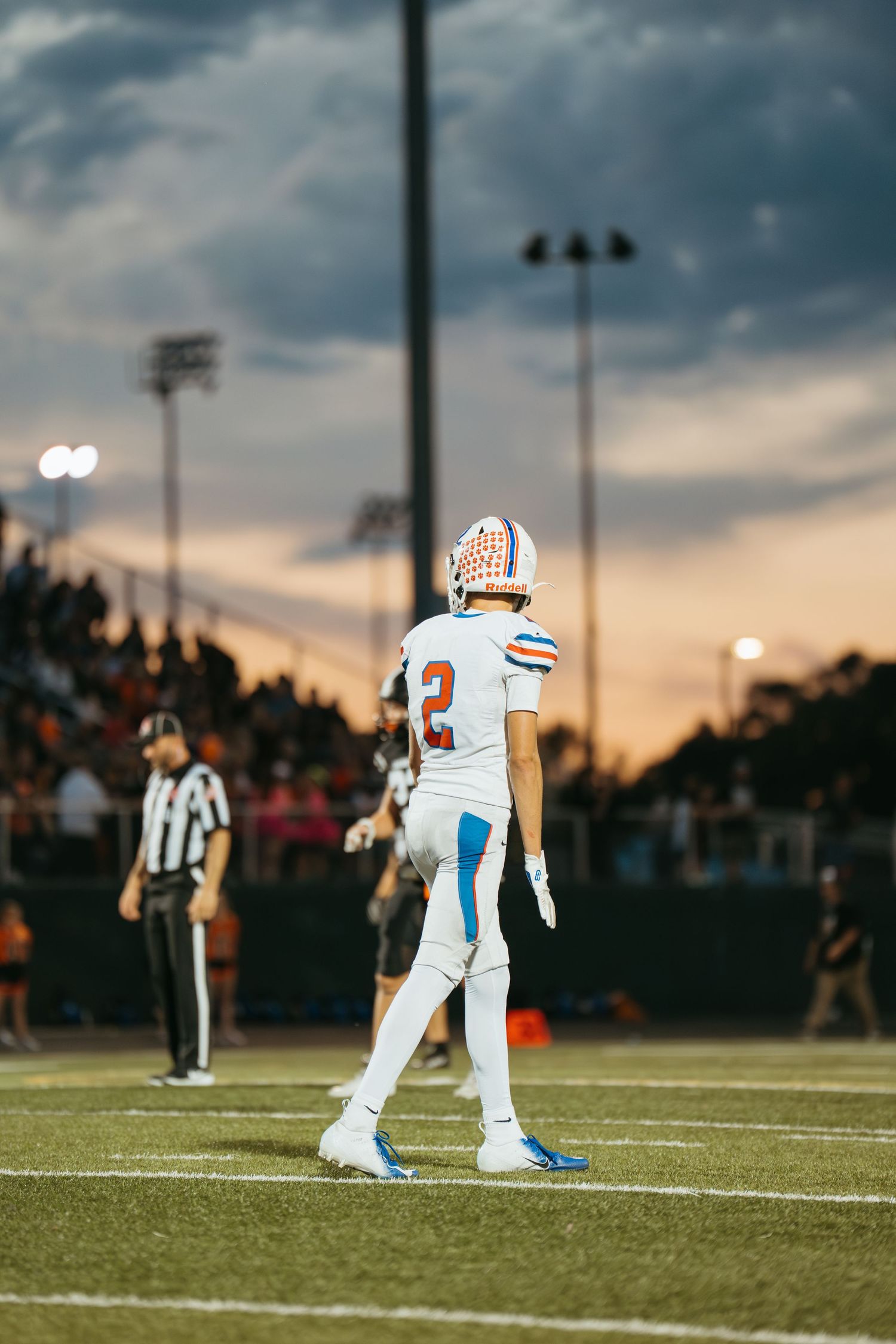 Football player wearing number 2 in white and orange uniform stands on field at dusk during a high school game.
