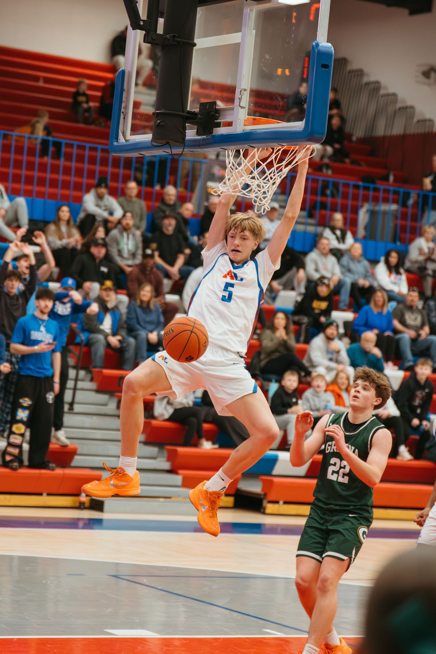 Player #5 in white dunks basketball while opponent #22 in green watches during high school basketball game.