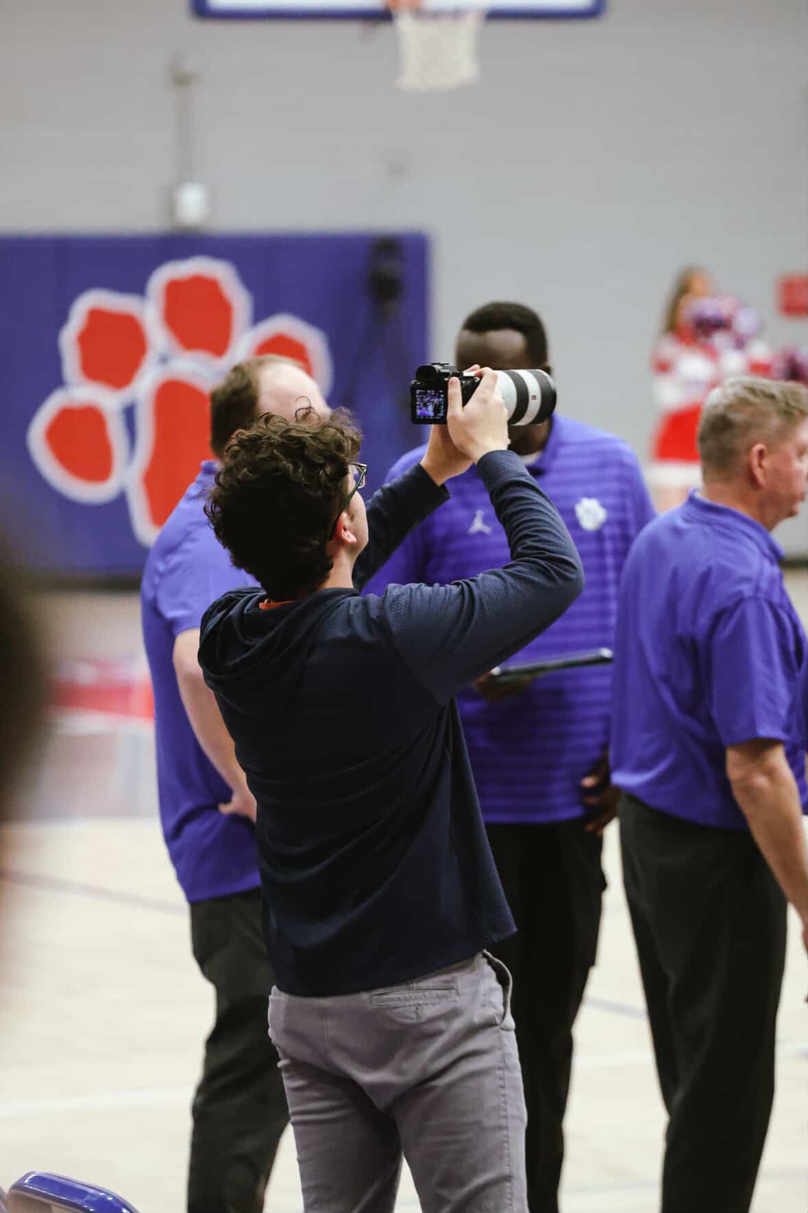 Photographer capturing footage at a basketball gym with players in purple Jordan Brand uniforms in the background.