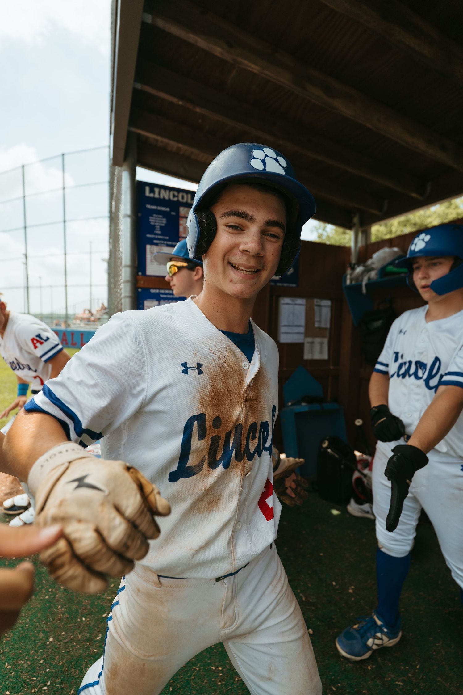 Smiling baseball player in Lincoln uniform with dirty gloves celebrates in dugout with teammates after scoring a run.