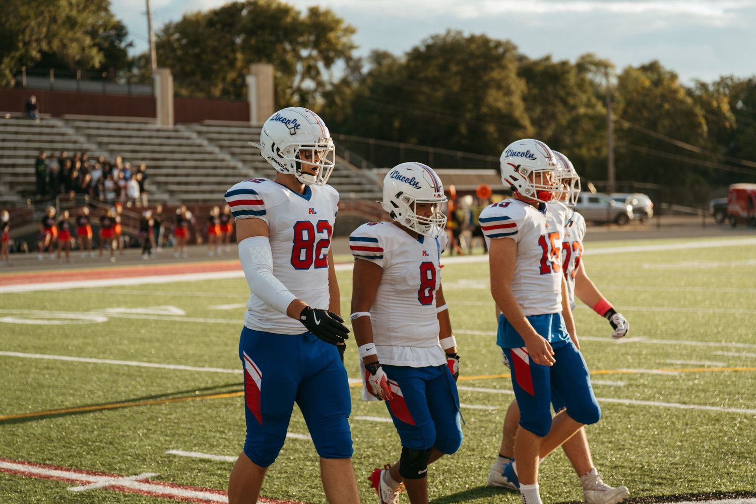 Three football players in white and red uniforms walk on the sideline during a game at an outdoor stadium.