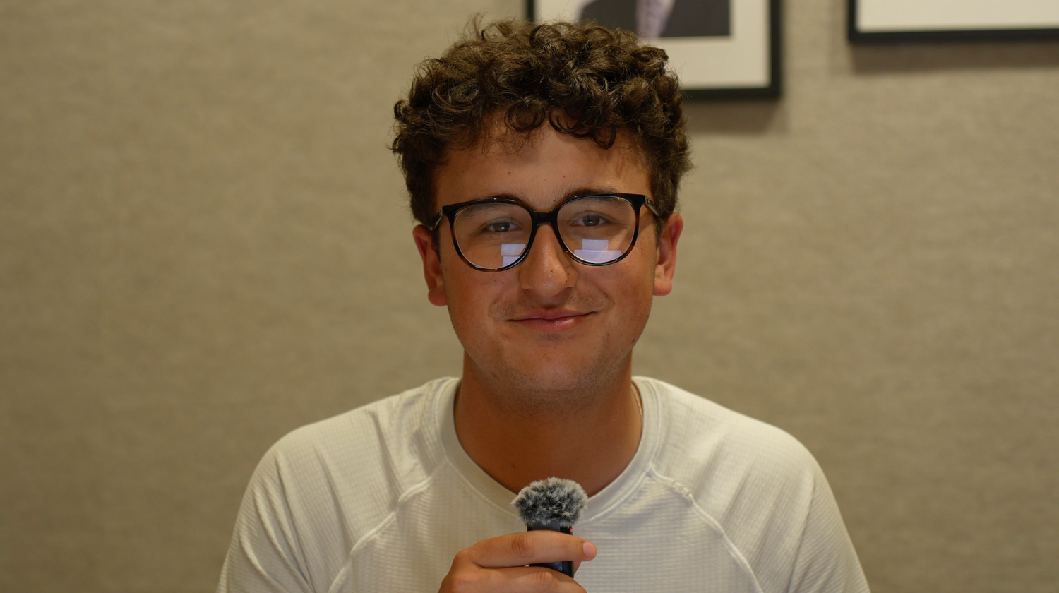 Young man with curly hair and black glasses holding a microphone, wearing a white t-shirt indoors.