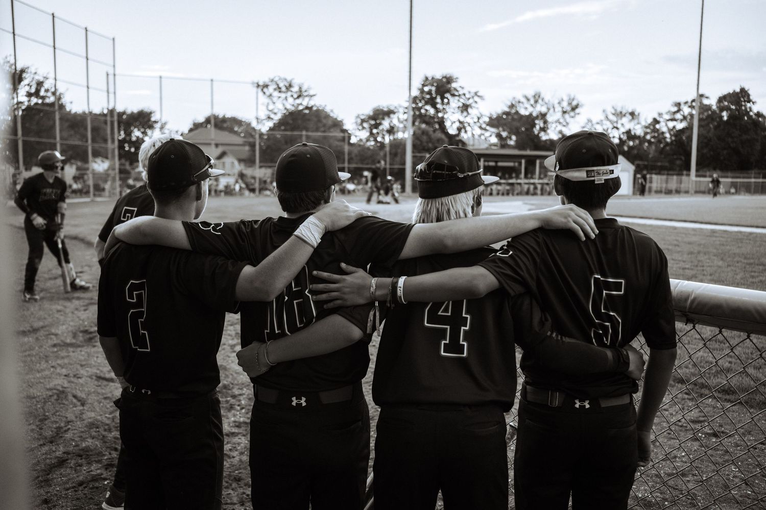 Four baseball players in black jerseys huddle together with arms around each other on a field.