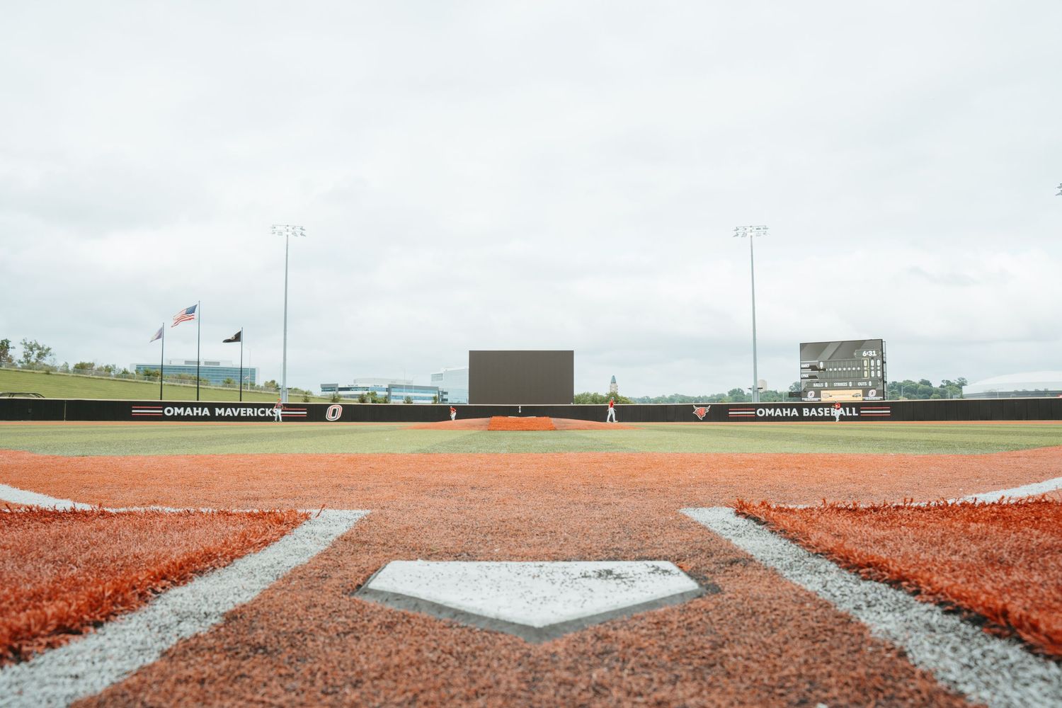 Omaha Mavericks baseball field view from home plate showing red clay diamond, green outfield, and scoreboard.