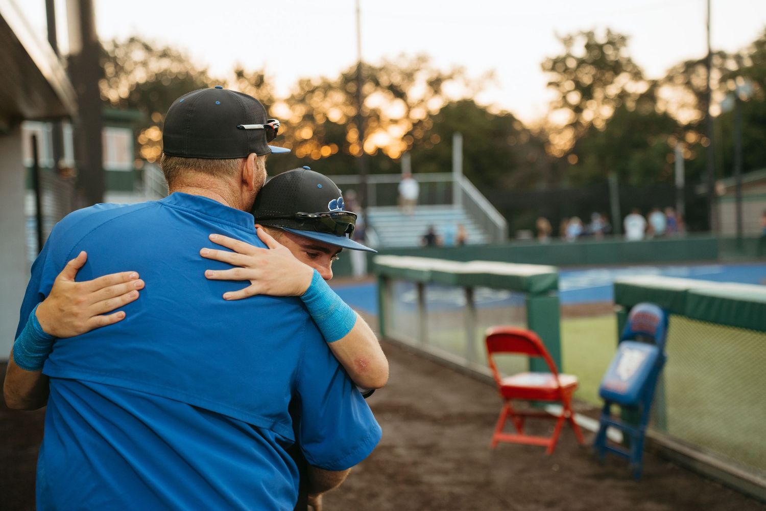 Coach and player share a heartfelt hug on a baseball field at sunset during a youth baseball game.