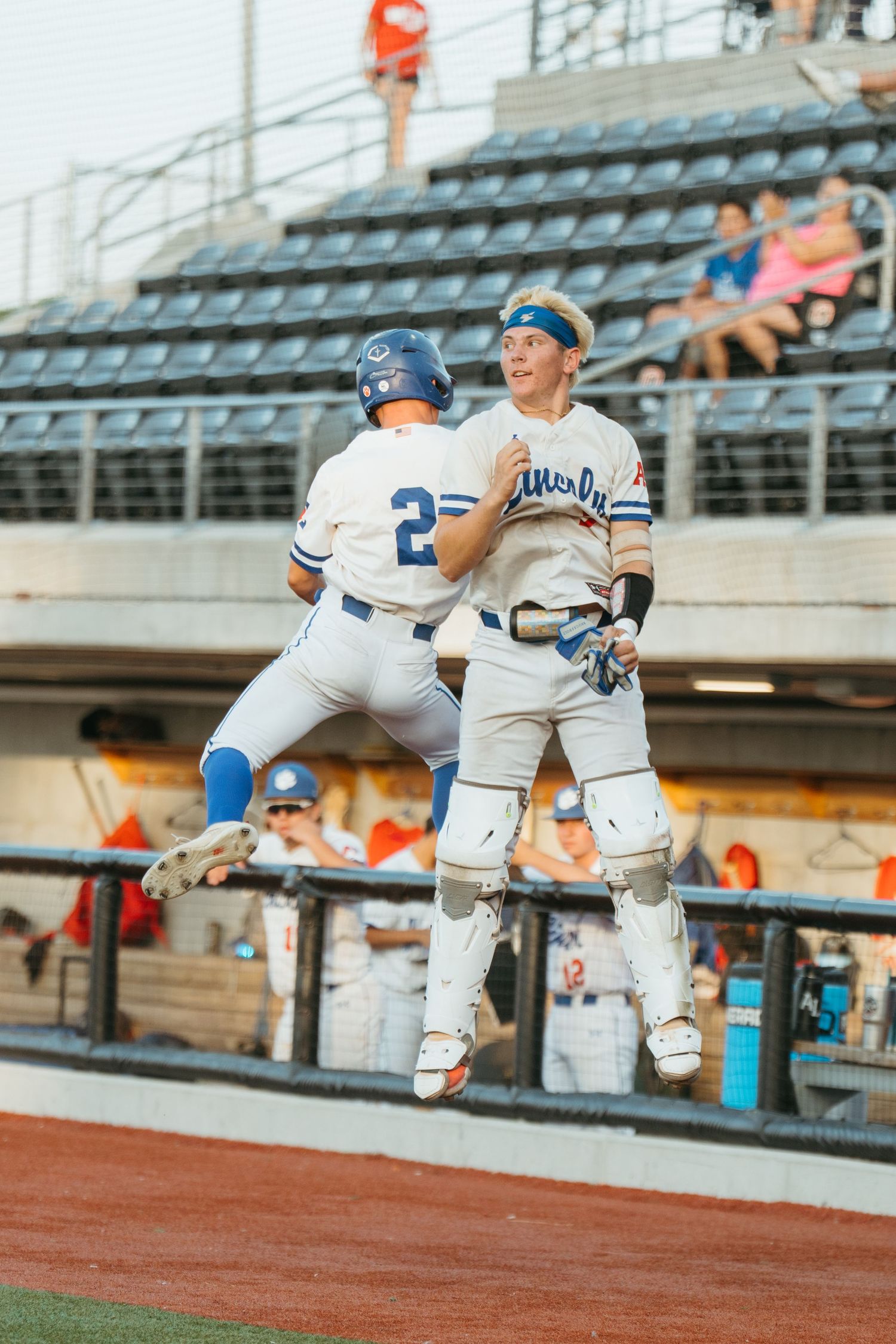 Two baseball players in white uniforms celebrate with a jump near the dugout during a game.