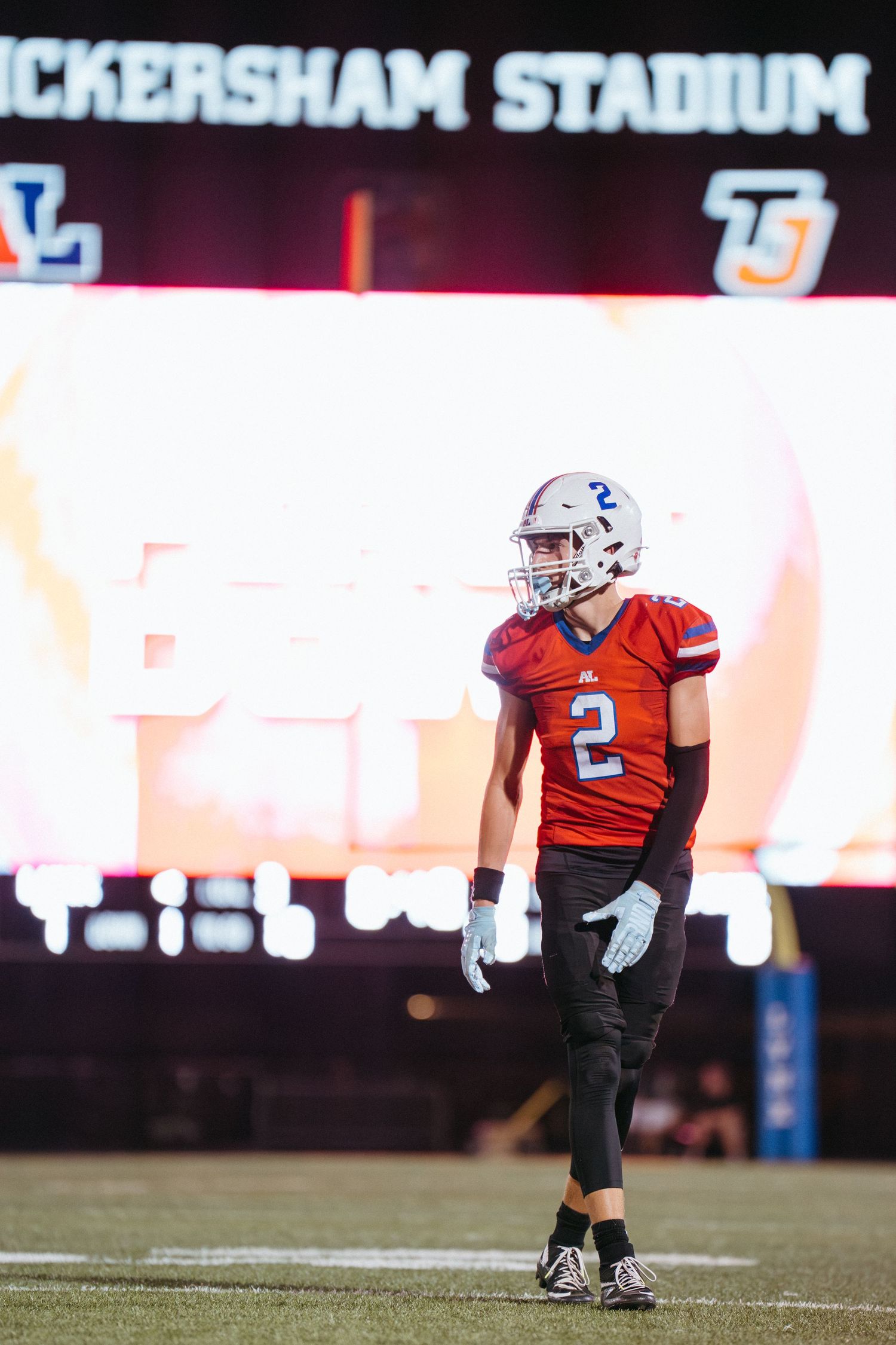 Football player wearing number 2 red jersey stands on field at Dickersham Stadium under bright lights.