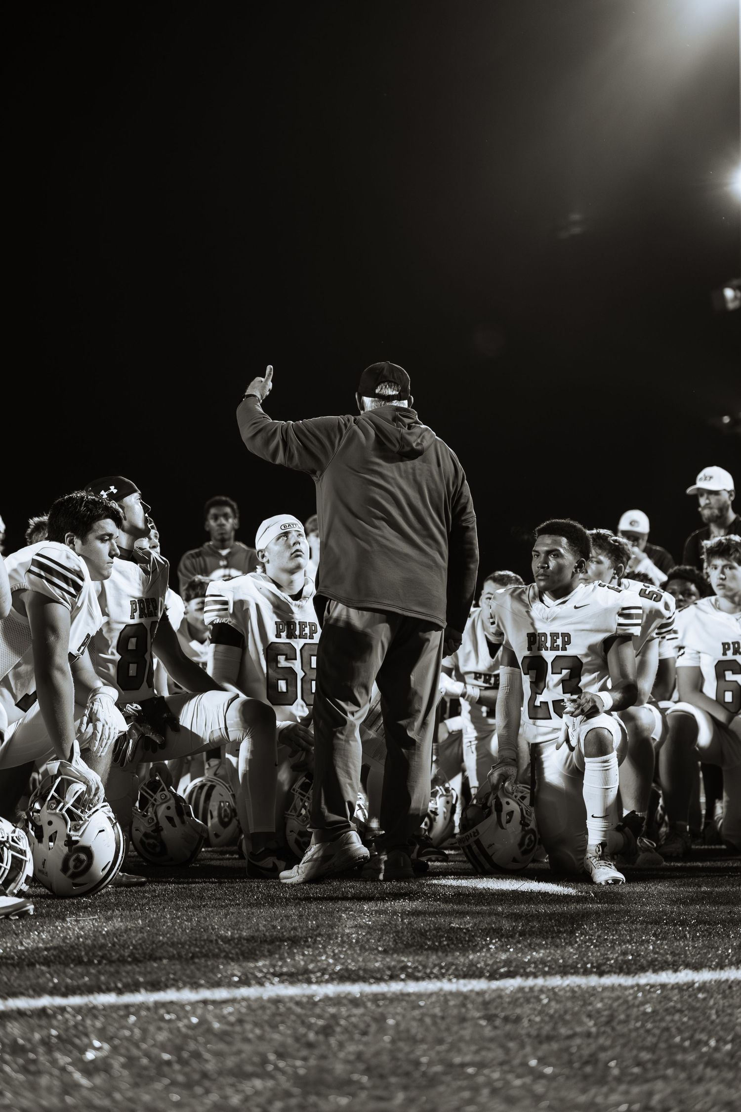 Black and white photo of a football coach addressing kneeling players on field at night after a game.