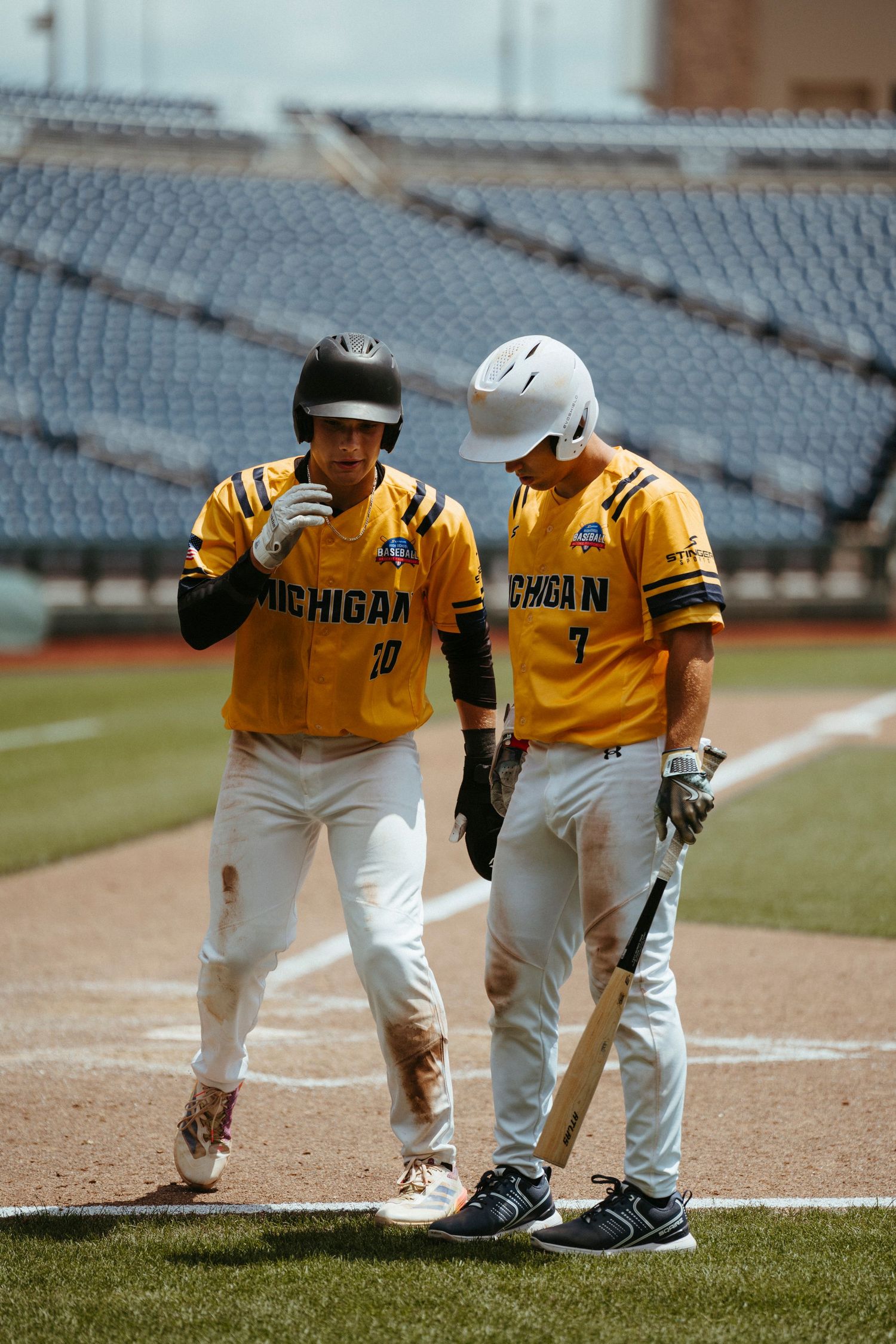 Two Michigan baseball players in gold uniforms discuss strategy on the field during a sunny game day.