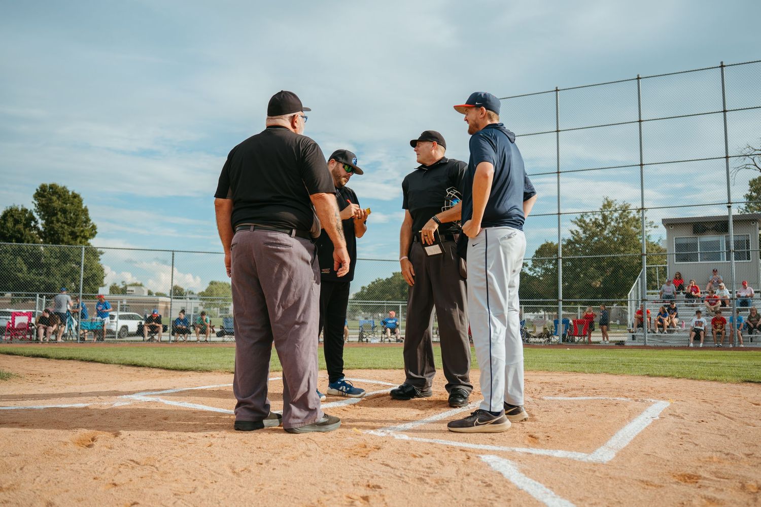 Baseball umpires and coaches meet at home plate before a game on a sunny day at a community baseball field.