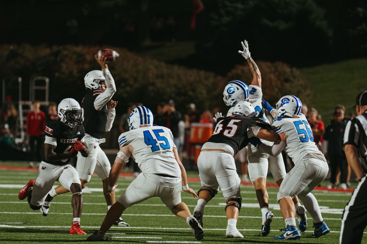 Football quarterback throws a pass under pressure during a competitive night game under stadium lights.