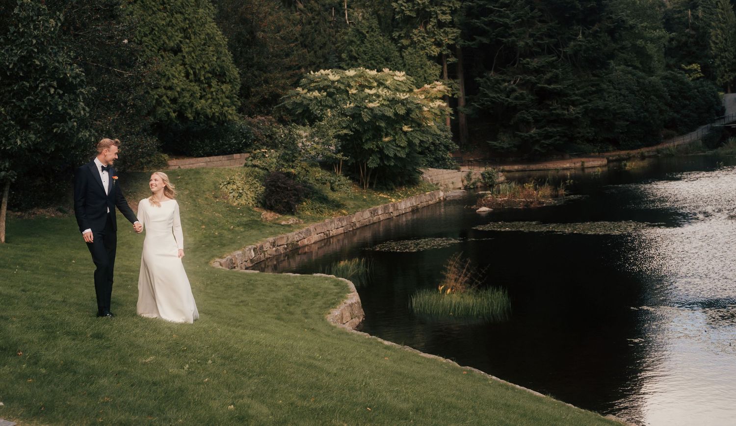 A couple poses together on a grassy lawn near a serene pond during their wedding day photo session.