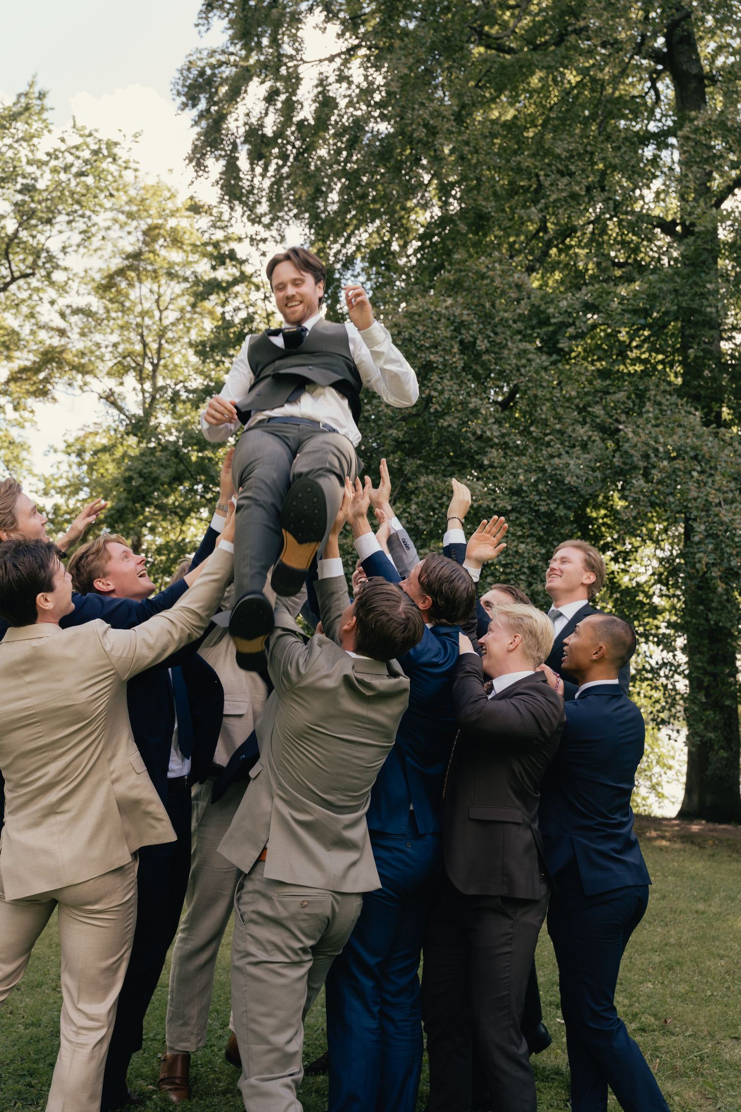 Groomsmen lifting groom into the air in celebration outdoors surrounded by trees at wedding reception.