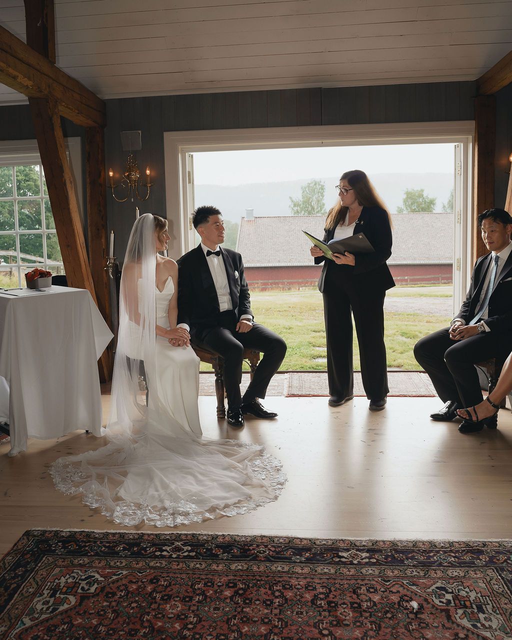 Wedding party members sit and kneel on wooden floor in rustic venue with oriental rug and natural lighting.