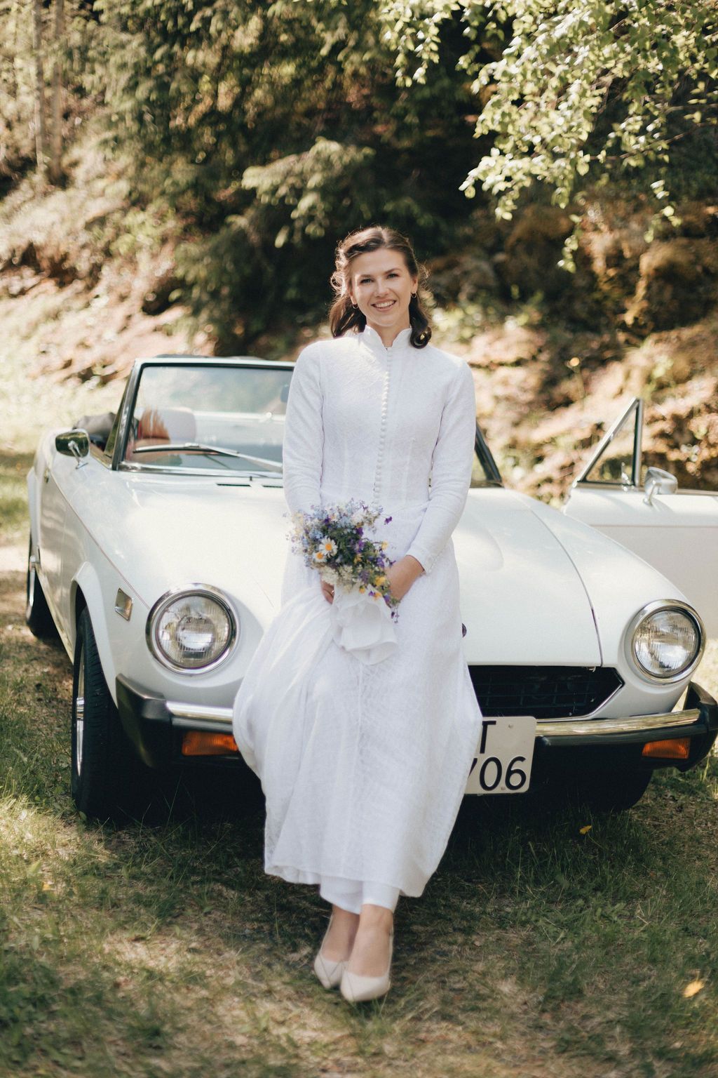 A vintage white convertible car parked in a garden setting with someone in an elegant white dress holding a bouquet.
