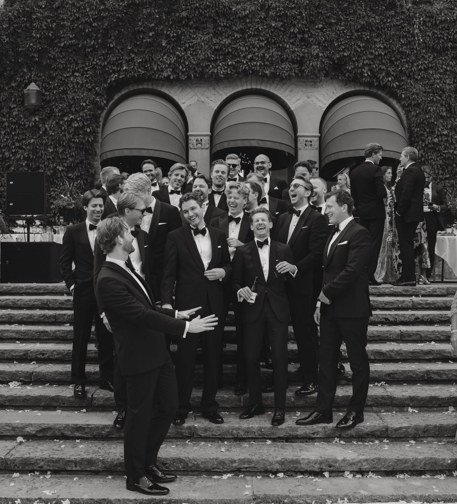 Black and white photo of laughing groomsmen in tuxedos gathered on stone steps in front of an ivy-covered building.
