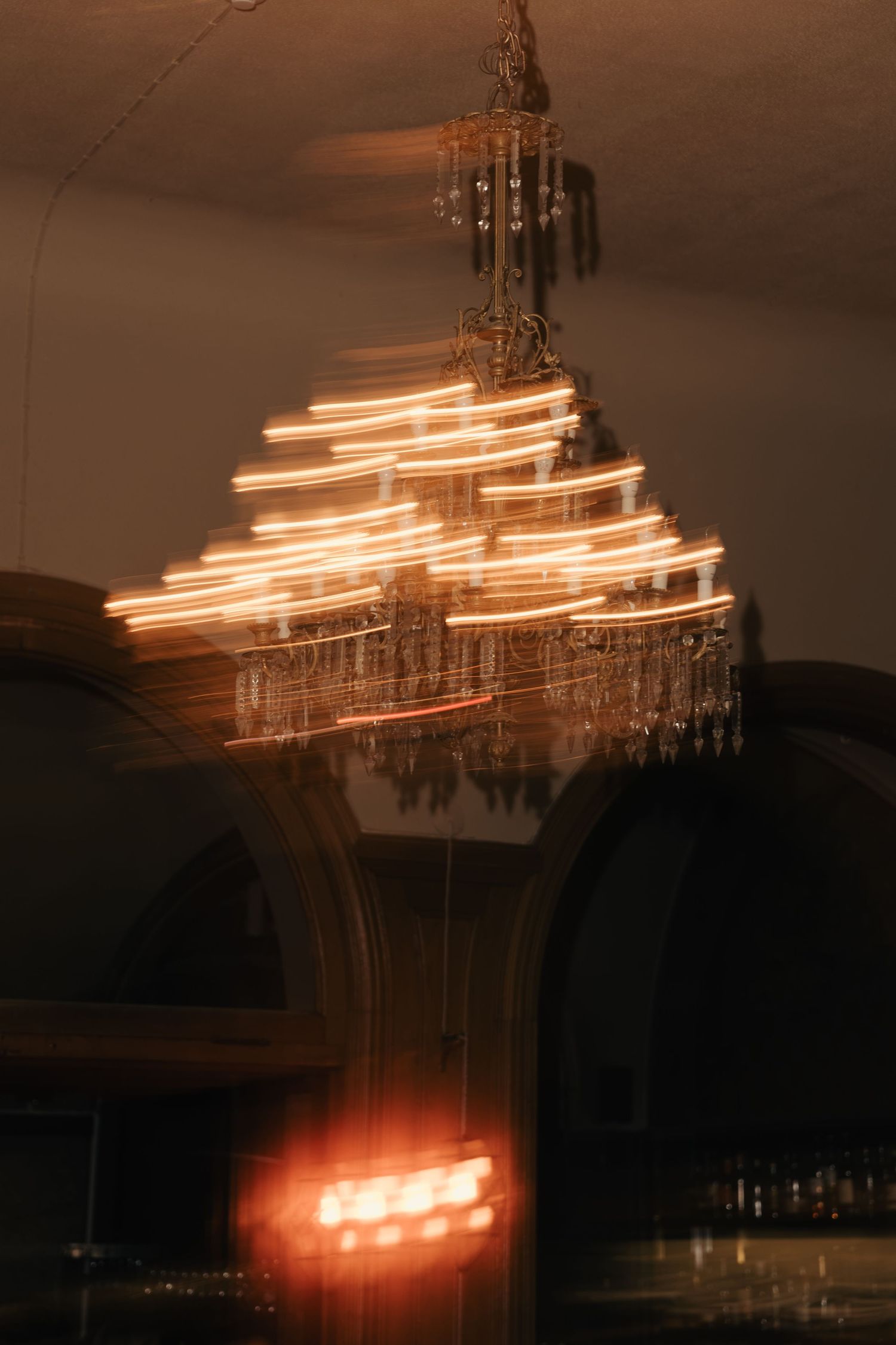 Long-exposure photo of an ornate crystal chandelier with glowing light trails in a dimly lit room with arched doorways.