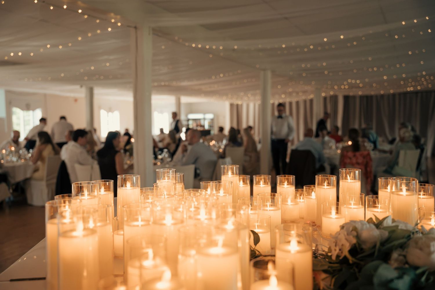 Glowing white pillar candles in glass holders light an elegant wedding reception hall with draped ceilings and guests seated at tables.