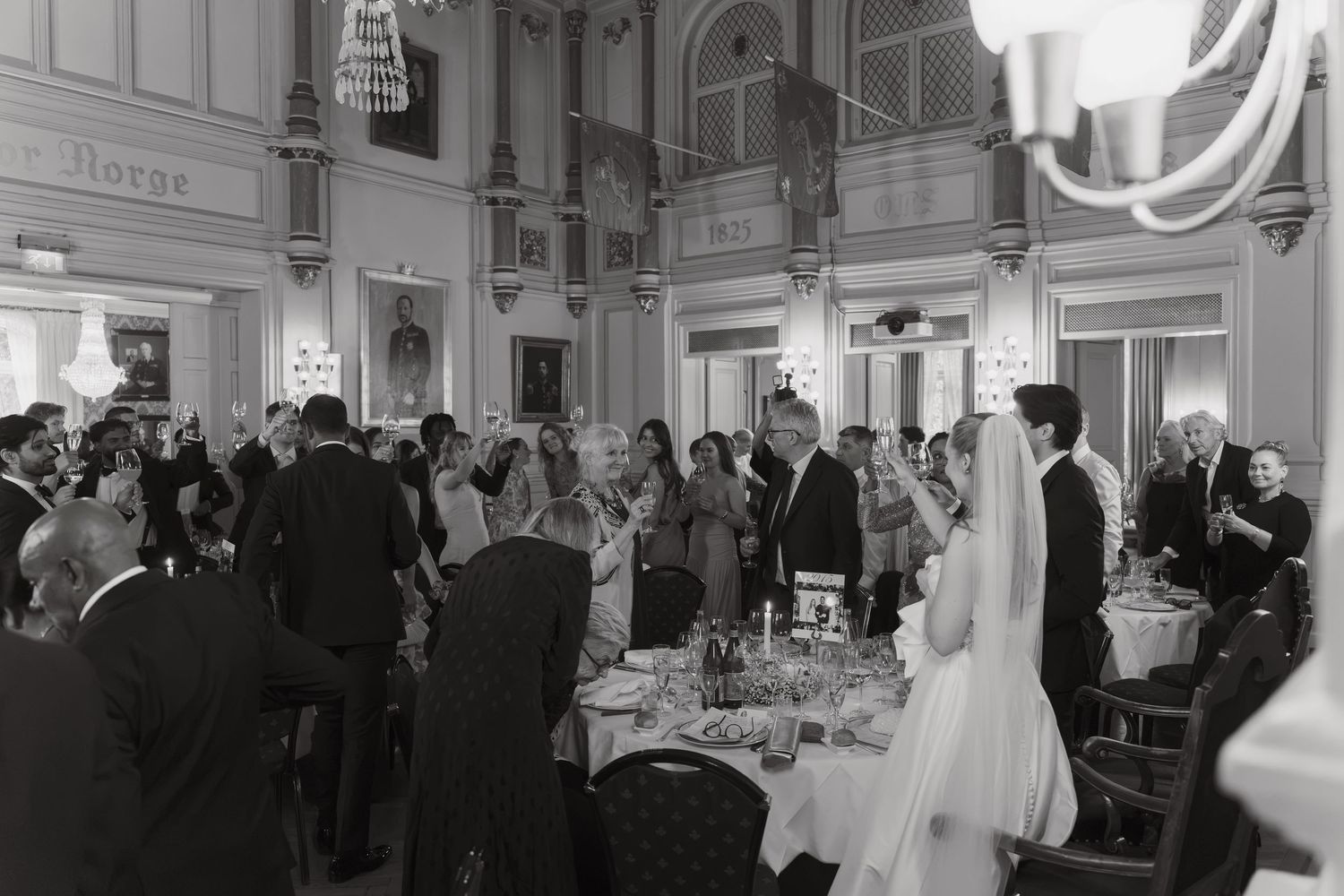 Black and white photo of wedding guests gathered around dinner tables in an elegant ballroom as the bride stands nearby.