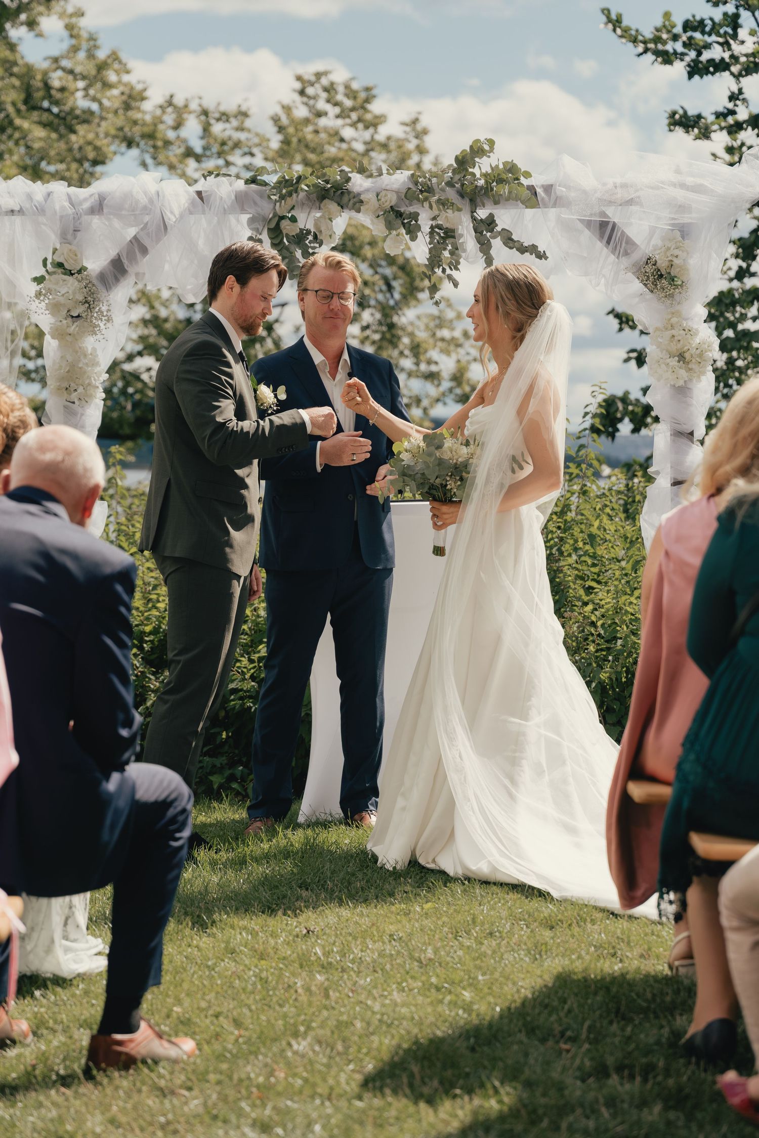 Bride and groom exchange rings at outdoor wedding altar decorated with white draping and florals.