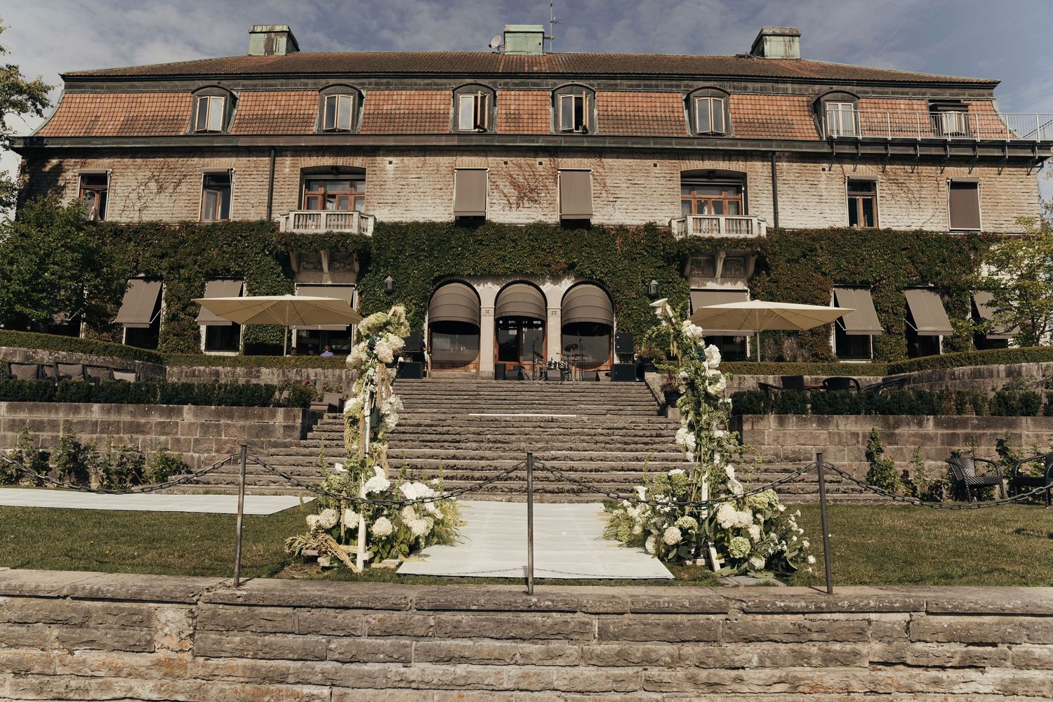 Elegant stone manor with ivy-covered terraces and a wide staircase flanked by white floral arrangements, ideal for a scenic wedding venue.