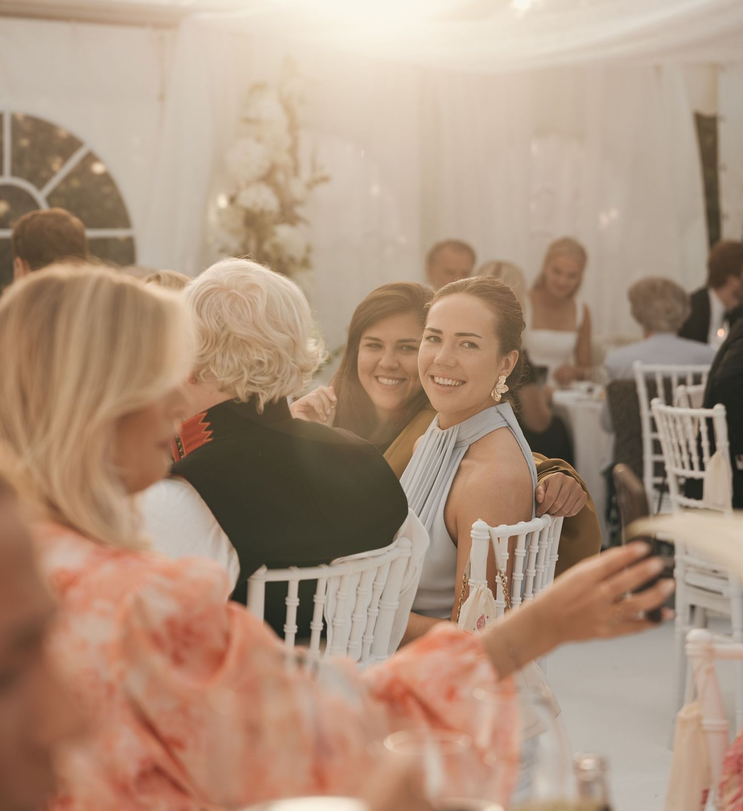 Smiling woman in a light blue dress sits among laughing wedding guests at a reception inside an elegant white tent.