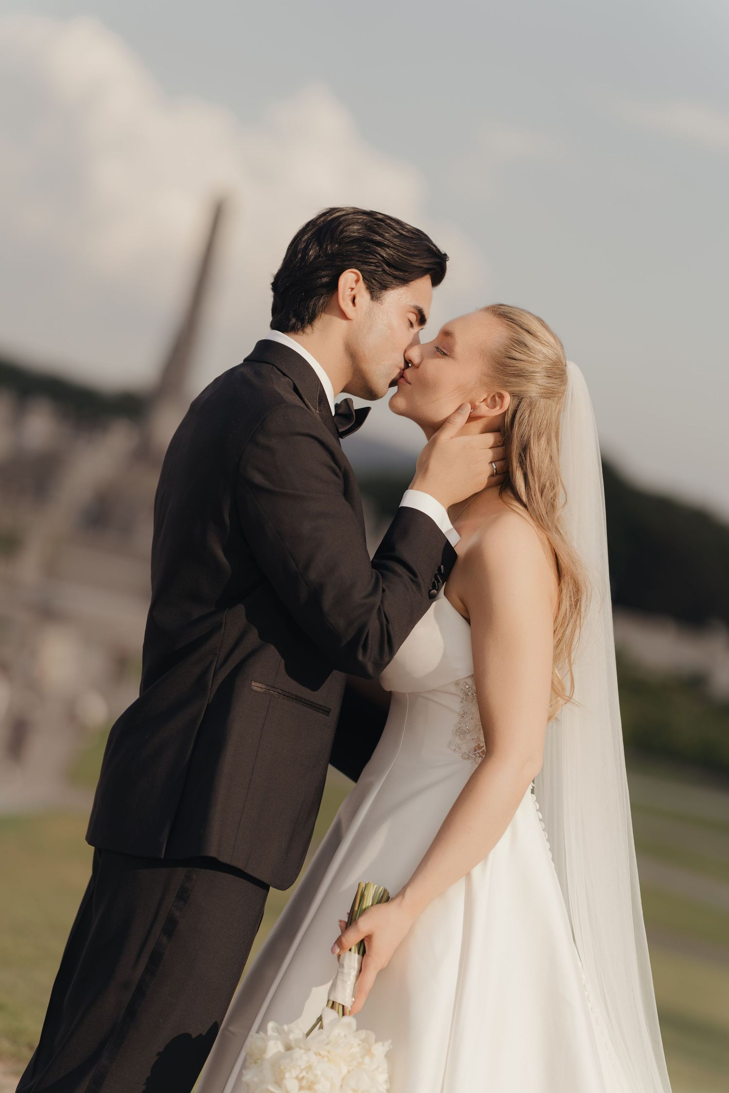 Groom in a black tuxedo and bride in a white gown with veil share a romantic kiss outdoors on their wedding day.