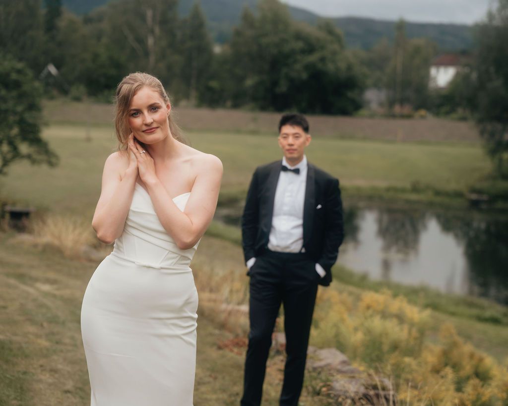 A bride and groom pose together in an outdoor setting with mountains, trees and a lake in the background.