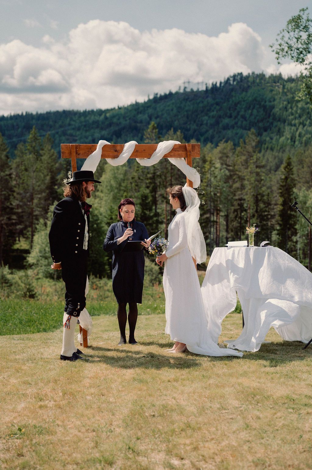 A rustic outdoor wedding ceremony takes place in a mountain meadow with evergreen forests and cloudy skies in the background.