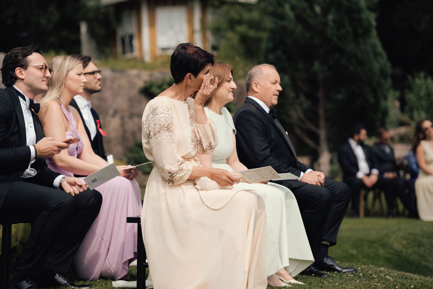 Guests seated outdoors at a wedding ceremony, with a woman in lace dress wiping tears during emotional moment.