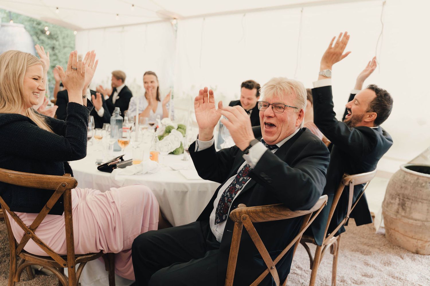 Wedding guests clapping and raising hands in celebration at a reception dinner inside a white marquee tent.