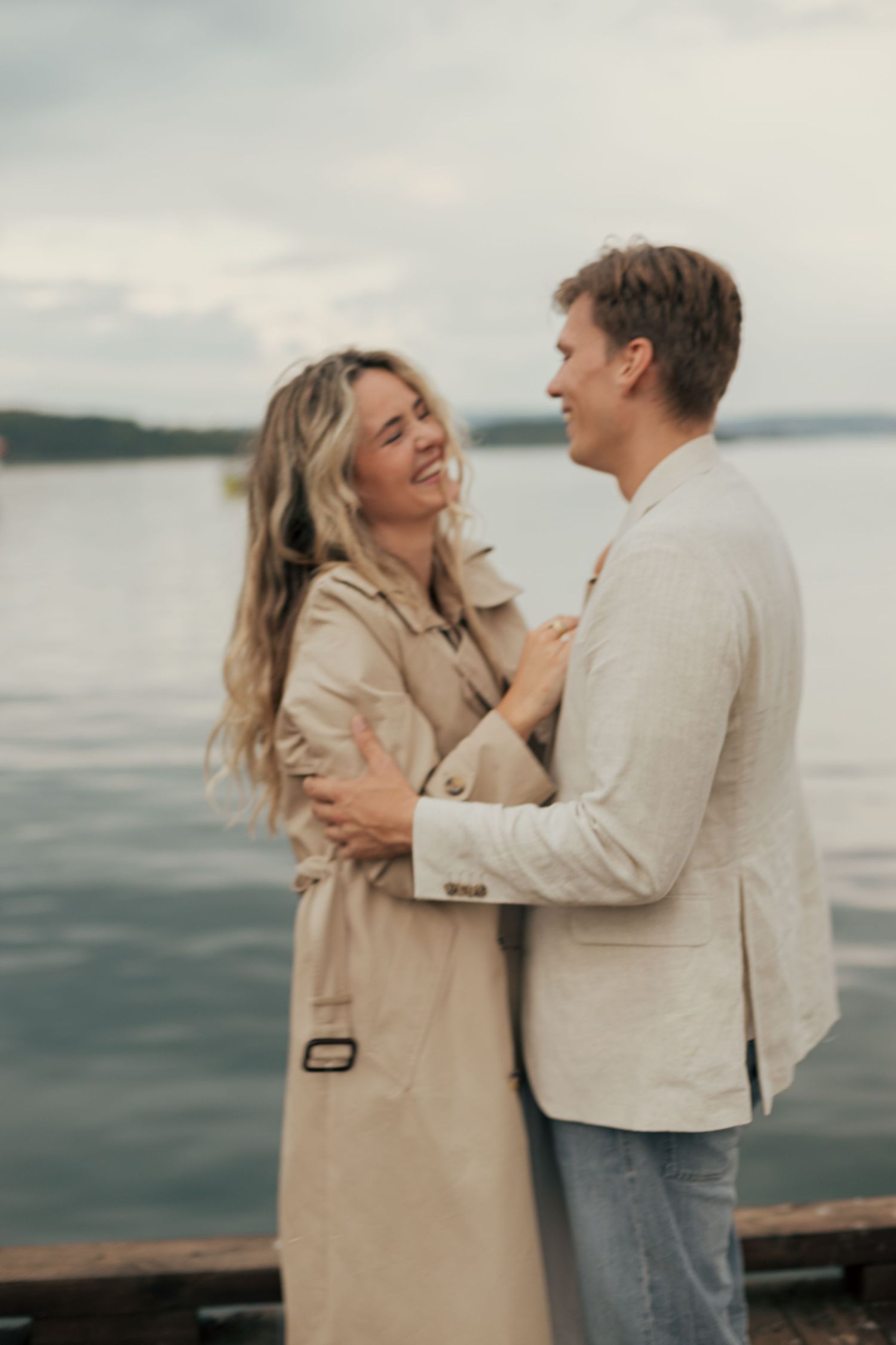 A couple shares joyful moments together while standing near a waterfront on an overcast day.