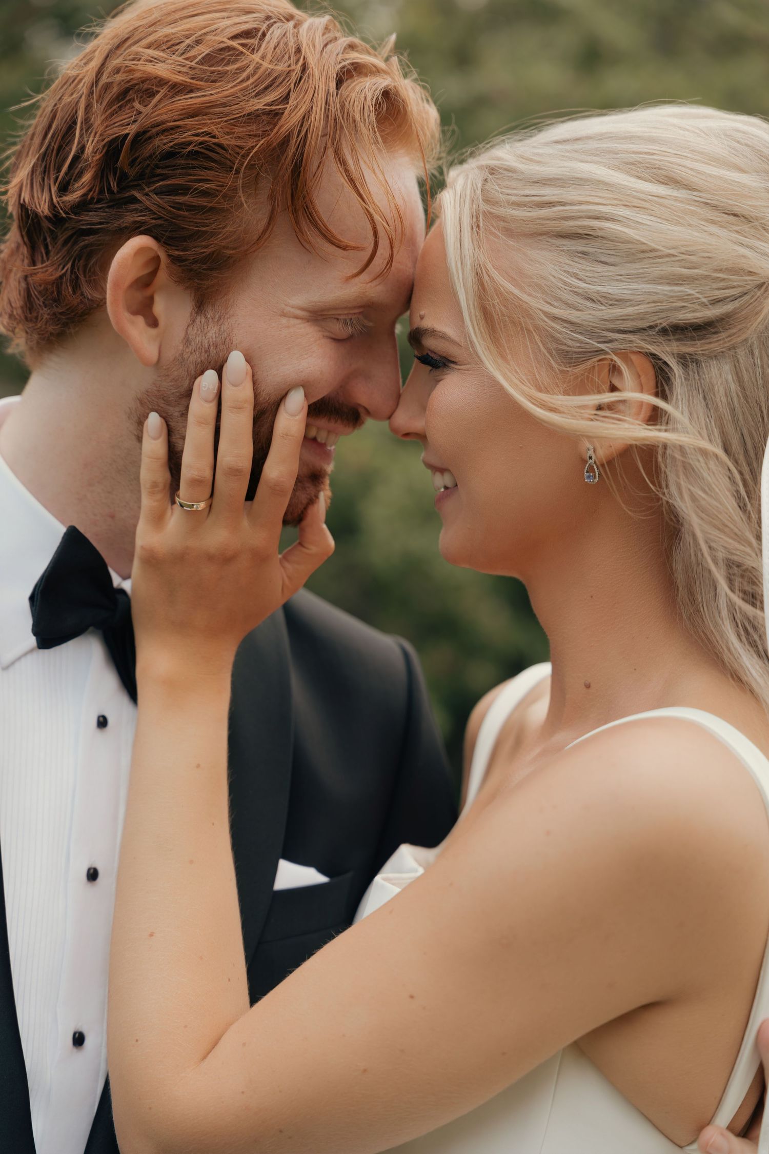 Smiling bride in a white dress touches her groom's face as they press foreheads together in a tender outdoor wedding portrait.