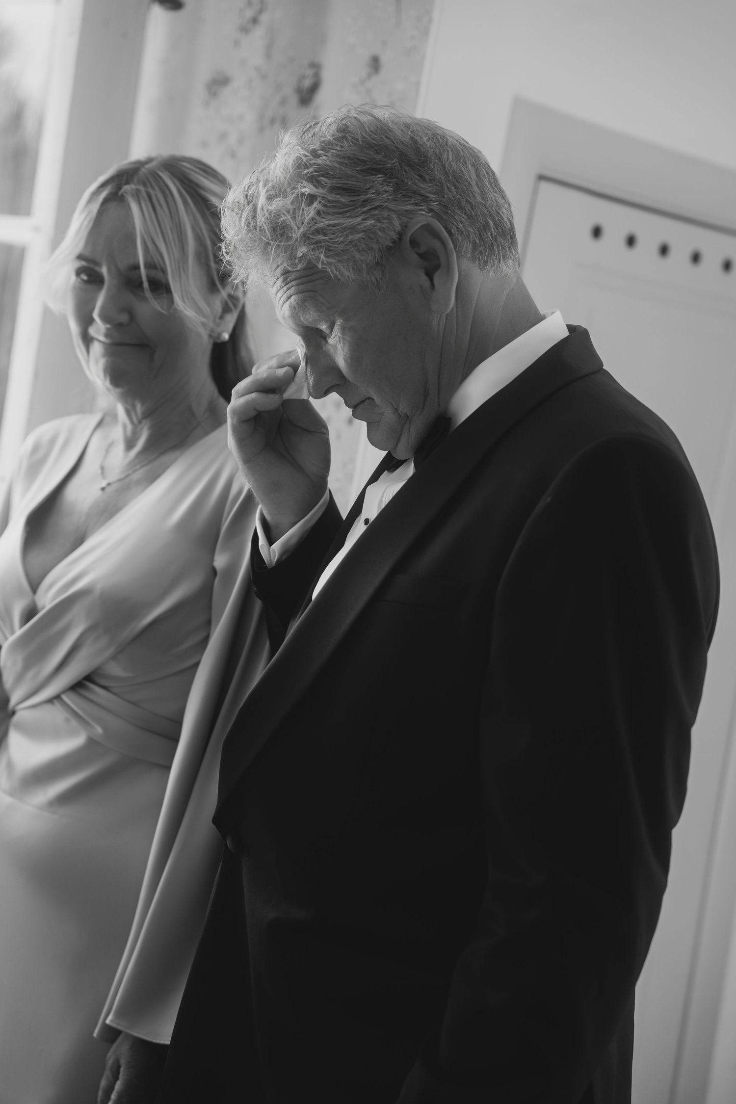 Black and white photo of an emotional man in a dark suit wiping his eye as a smiling woman in a draped dress stands beside him.