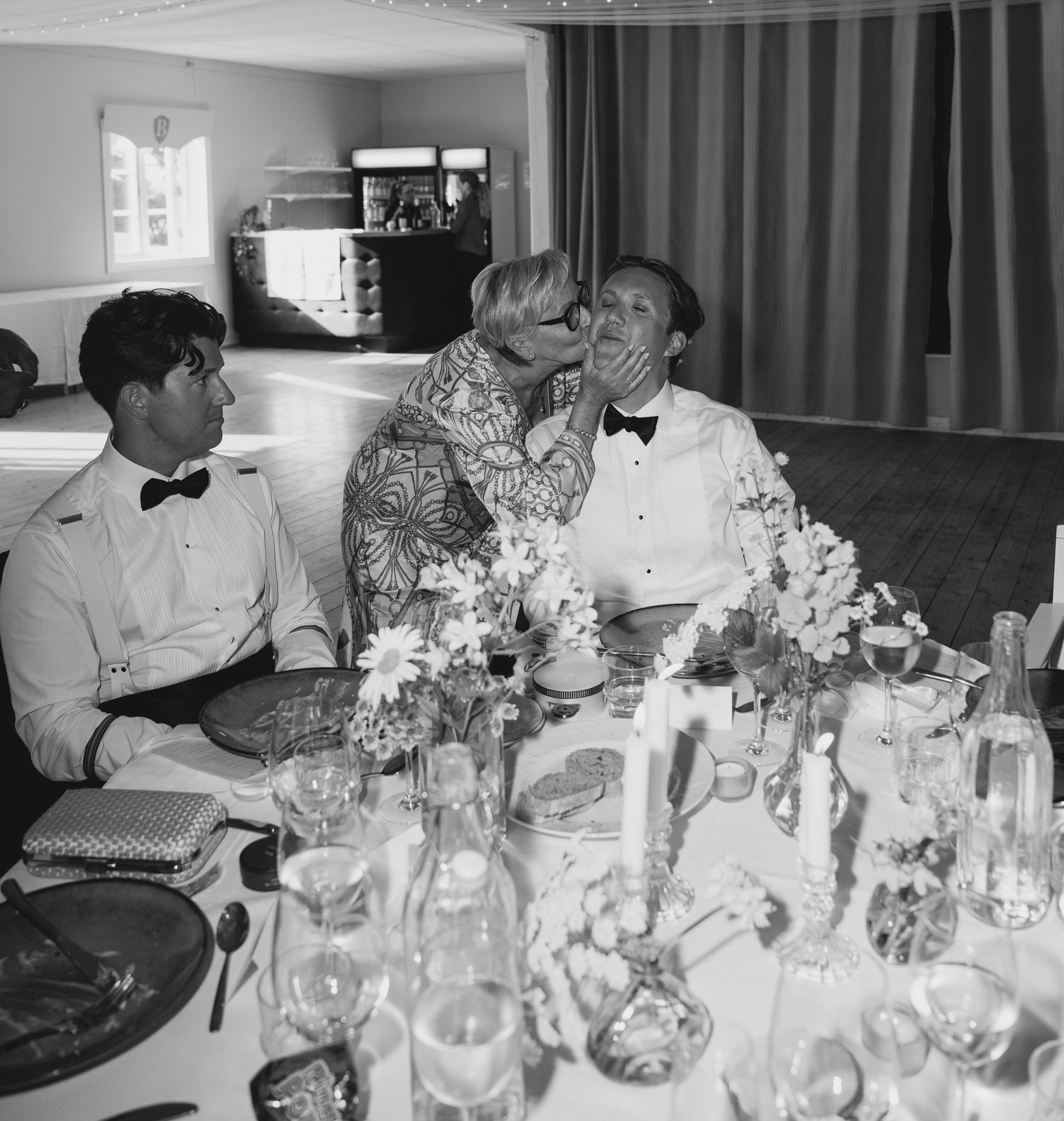 Black and white wedding reception photo of a woman kissing a groom on the cheek beside a seated man at a table with flowers and candles.