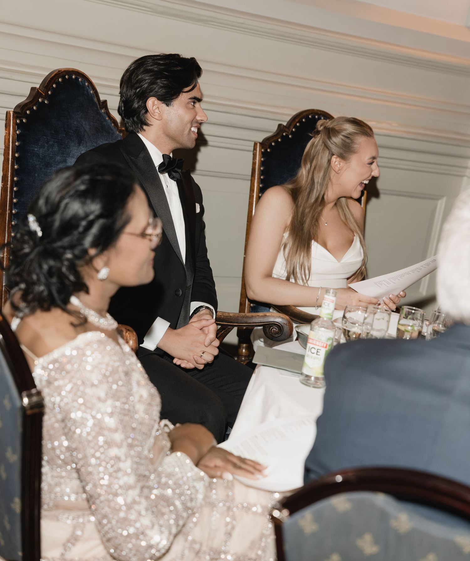 Groom in a tuxedo and bride in a white dress smile from ornate chairs at a wedding reception while a guest in sequins looks on.
