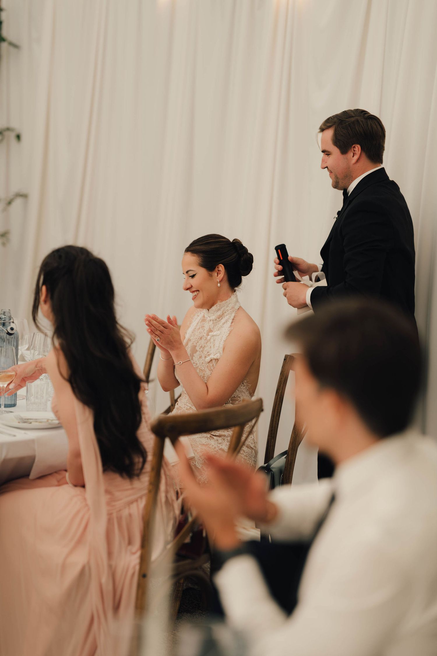Bride smiling and clapping at wedding reception while groom stands giving a toast in elegant venue.