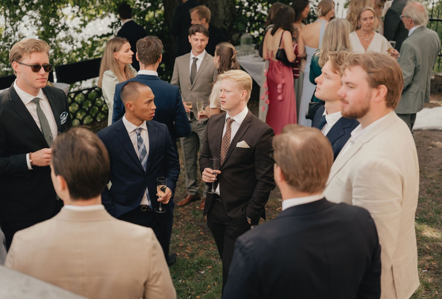 Elegantly dressed guests mingling outdoors at a wedding reception, wearing suits and formal attire.