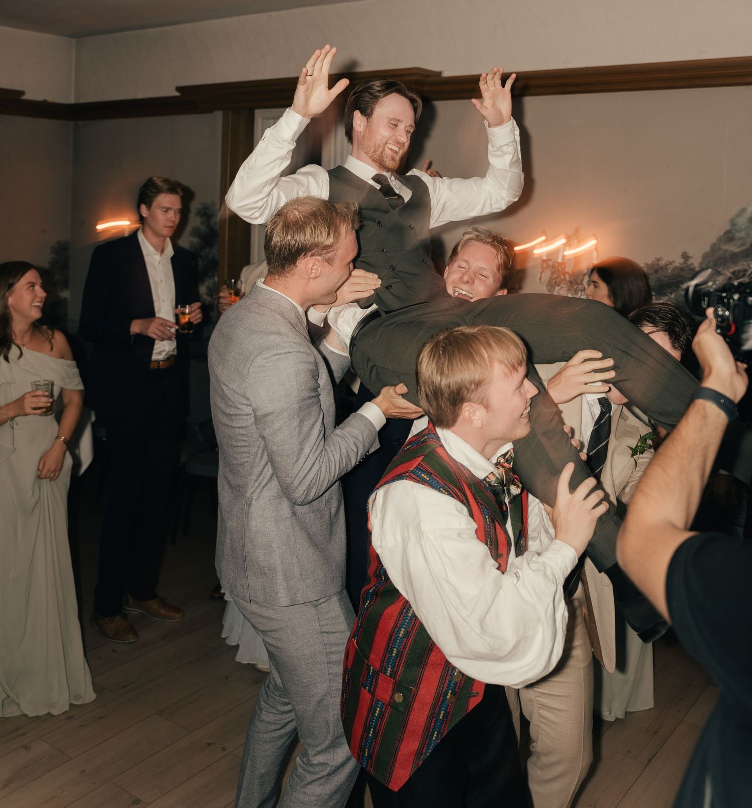 Groomsmen lifting groom in the air during a lively wedding reception celebration indoors.