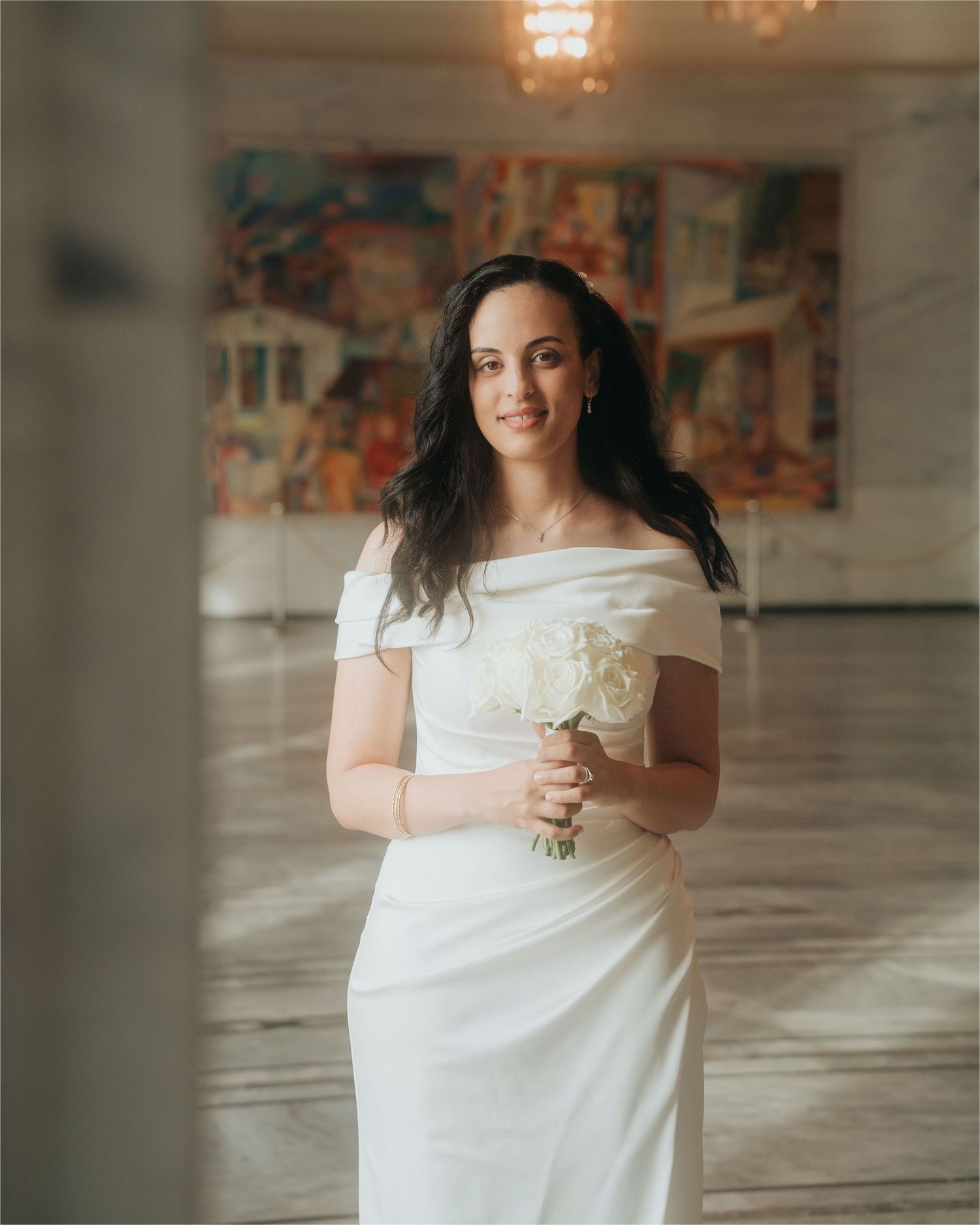 A bride in a white off-shoulder gown poses gracefully in a light-filled room with elegant chandeliers and abstract art.