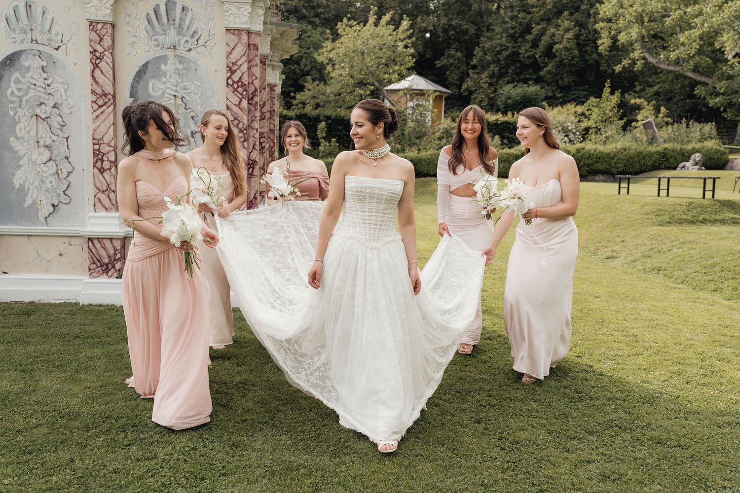 Bride in white gown walks with four bridesmaids in blush pink dresses carrying bouquets in a lush garden setting.