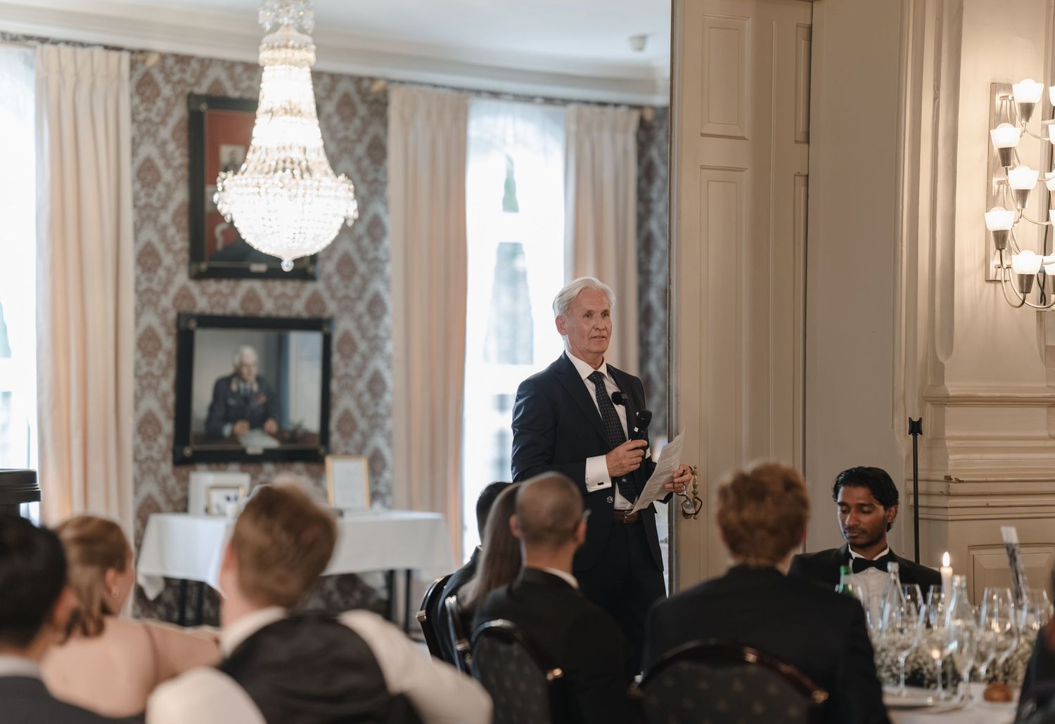 Man in a dark suit gives a speech to seated guests at an elegant banquet room with a crystal chandelier and framed portraits.
