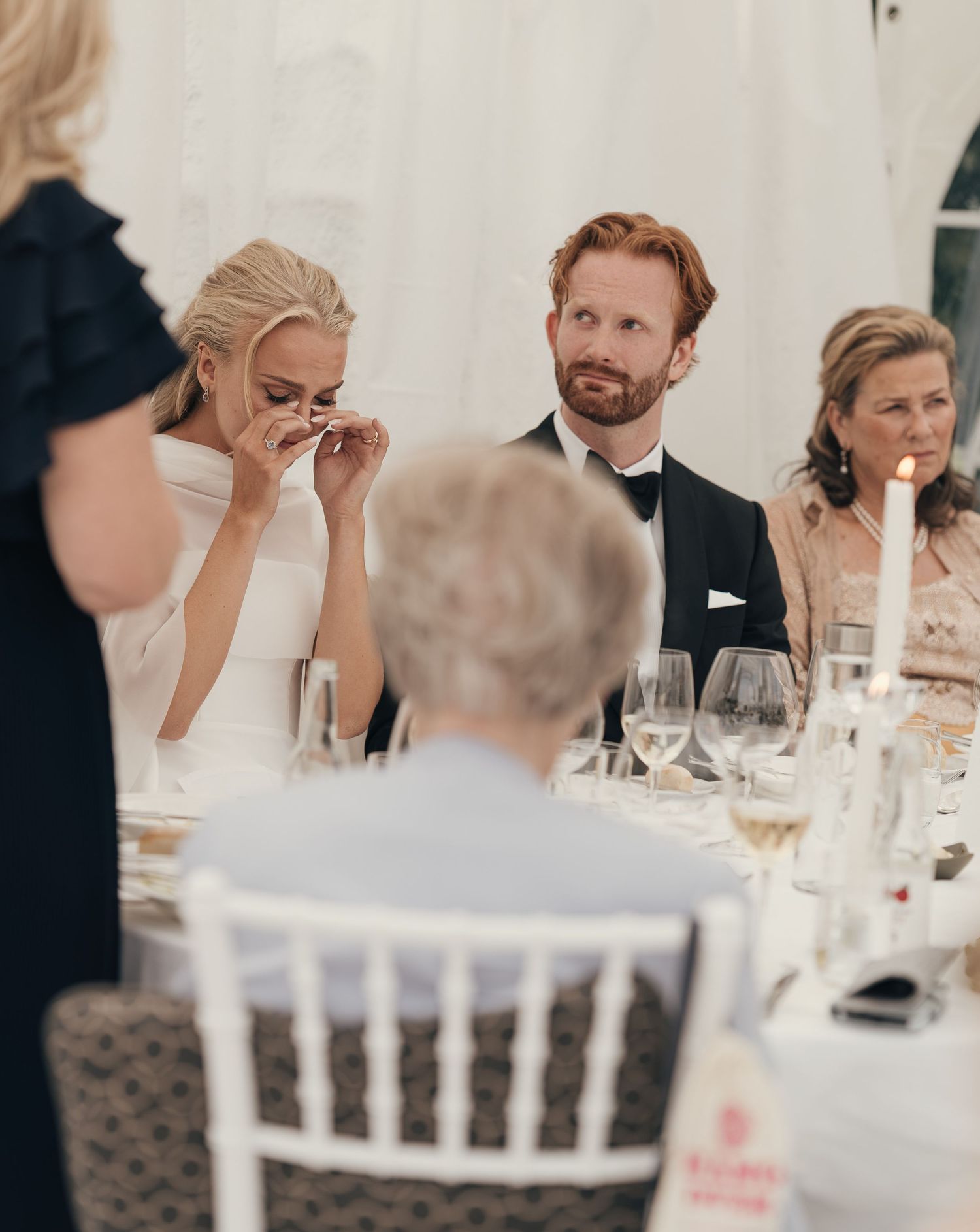 Bride wipes away tears of joy at the reception table beside the groom and guests during a wedding toast.