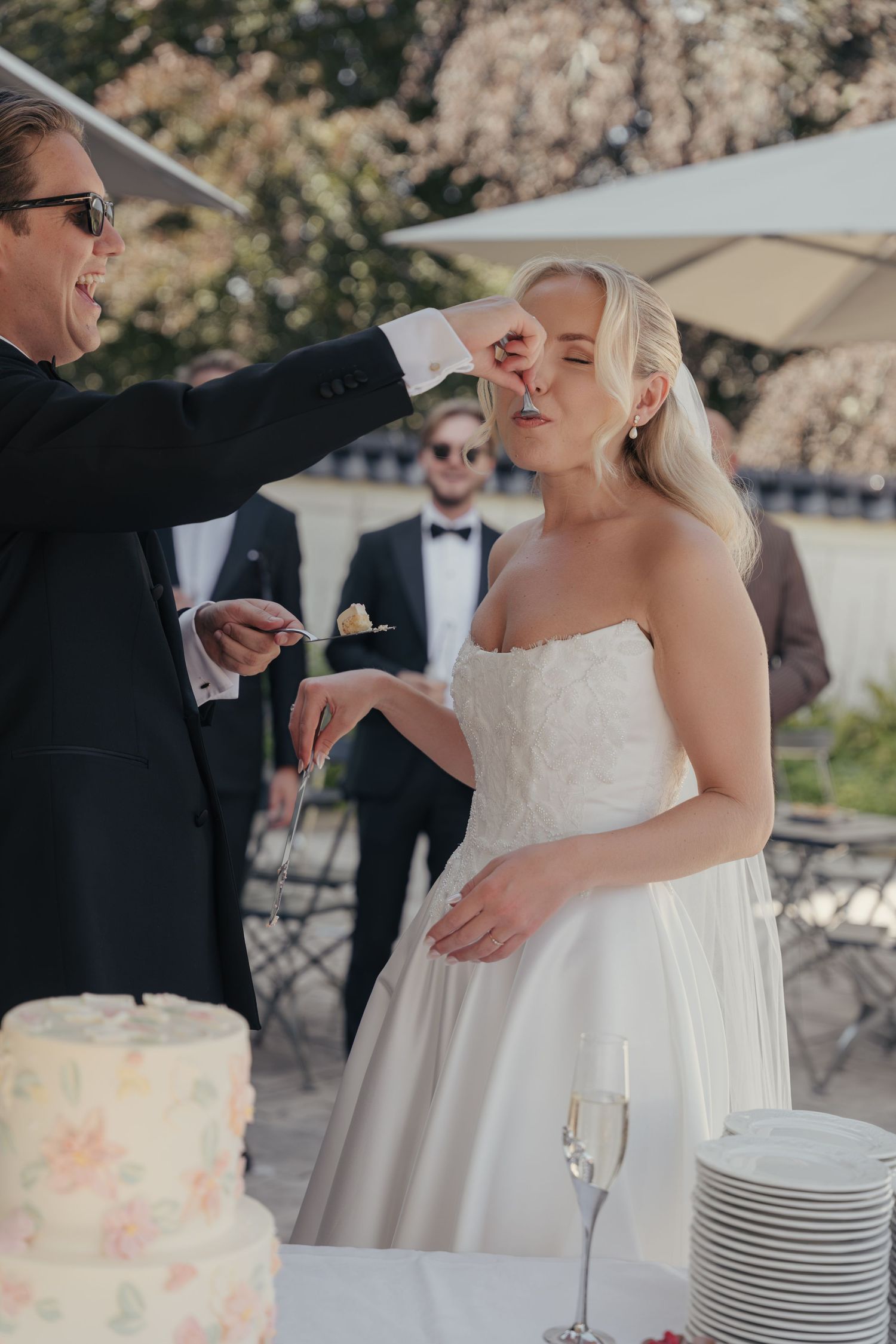 A bride in a strapless white gown tastes wedding cake fed by a groom in sunglasses at an outdoor garden reception.